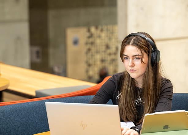 A student with headphones on studying in front of a laptop and a tablet device