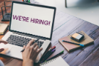 A laptop on a desk with scattered stationary. The text on the screen reads 'WE'RE HIRING!' 