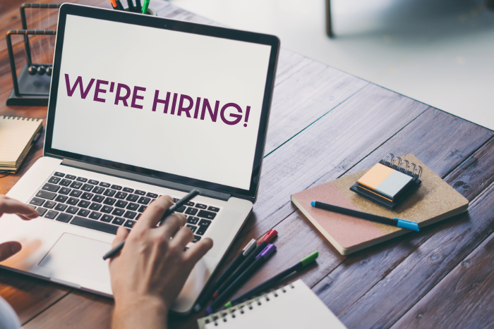 A laptop on a desk with scattered stationary. The text on the screen reads 'WE'RE HIRING!'