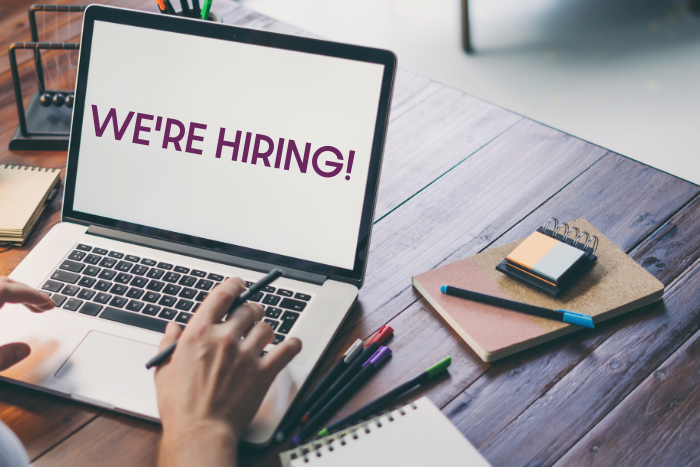 A laptop on a desk with scattered stationary. The text on the screen reads 'WE'RE HIRING!' 