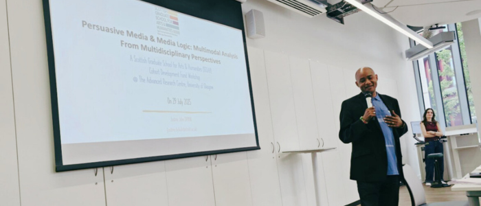 A speaker presents in a modern classroom with a large screen behind him displaying the workshop title “Persuasive Media & Media Logic: Multimodal Analysis From Multidisciplinary Perspectives” at the University of Glasgow