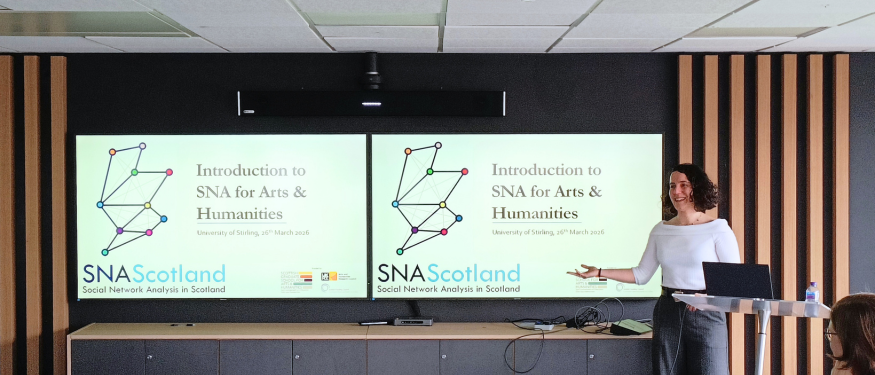 Presenter stands beside a lectern in a lecture room, pointing at two screens displaying slides titled “Introduction to SNA for Arts & Humanities” with network diagrams and the SNA Scotland logo
