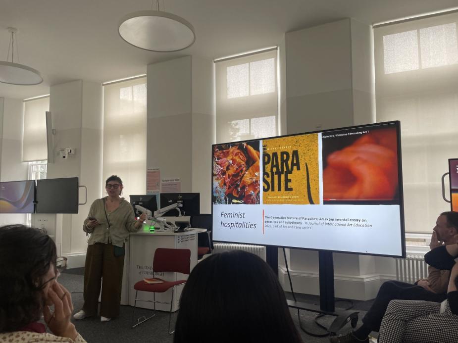 A presenter stands at the front of a university seminar room beside a large screen displaying a slide titled “Feminist hospitalities,” while attendees sit and listen.