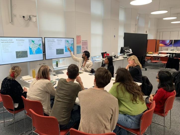 Participants seated around a table in a workshop setting, listening to a presenter who points to slides displayed on two large screens.