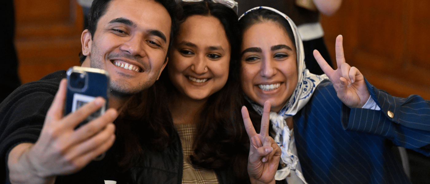 Three students posing for a selfie indoors, with two making peace signs while looking towards a smartphone camera.