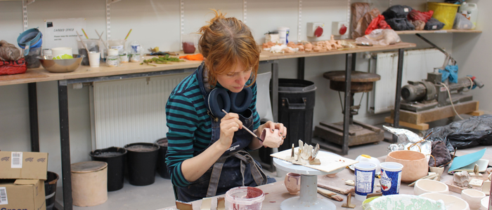 Research intern shaping a small clay sculpture at a workbench in a sculpture workshop, surrounded by tools, materials and ceramic pieces.