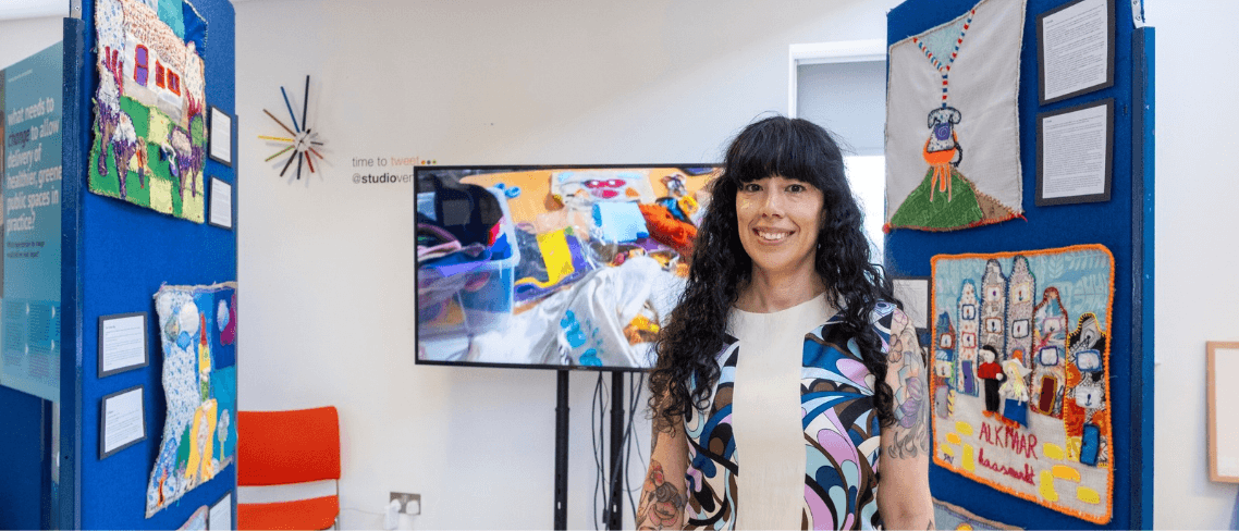 Doctoral researcher standing in an exhibition space surrounded by colourful textile artworks and a digital display.